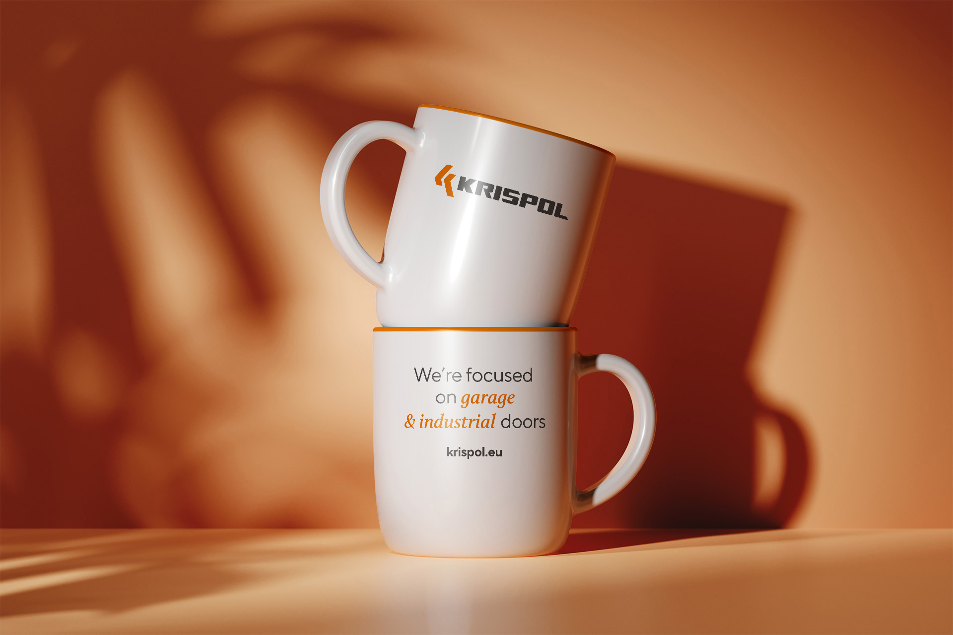 Obrazek przedstawia dwa kubki z logo firmy Krispol. Jeden kubek jest ustawiony na drugim, a na obu widnieje hasło „We’re focused on garage & industrial doors” oraz adres strony internetowej. Kubki mają białą barwę z pomarańczowym wykończeniem. Tło jest w ciepłych, pomarańczowych odcieniach.