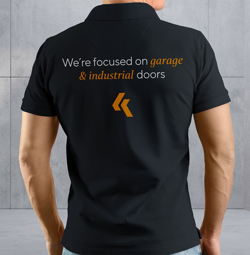 Na zdjęciu znajduje się czarna koszulka polo z napisem "We're focused on garage & industrial doors" na plecach.